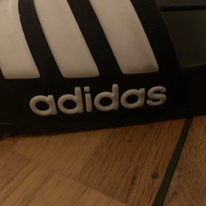 Svarta och vita sandaler från Adidas - Säljer ett par klassiska svarta sandaler från Adidas med de ikoniska tre vita ränderna över foten. Perfekta för sommarens varma dagar eller som bekväma inneskor. De har en enkel och stilren design med Adidas-loggan på sidan.