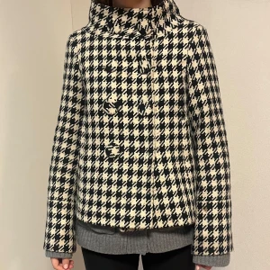 Rutig kappa med knappar - Snygg rutig kappa i svart och beige med stora knappar framtill. Jackan har en klassisk krage och långa ärmar, perfekt för höst och vår. Den har en stilren och tidlös design som passar till många olika outfits. Fodret inuti jacken är trasigt och en knapp saknas utanpå. Den är inte i toppskick men fortfarande fin💕