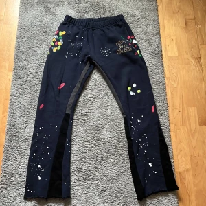 Mörkblå Gallery Dept byxor - Unika mörkblå byxor från Gallery Dept. med färgstänk i flera färger och textdetalj på sidan. Byxorna har en elastisk midja för extra komfort och en avslappnad passform. Perfekta för en kreativ och avslappnad stil. Byxorna passar folk mellan 172-181 cm.