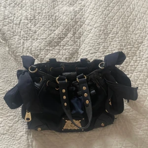 Mörkblå handväska från Juicy Couture - Snygg mörkblå handväska från Juicy Couture med gulddetaljer och knyt på sidorna. Väskan har flera fack med dragkedjor för säker förvaring. Perfekt för att bära med sig det viktigaste med stil.