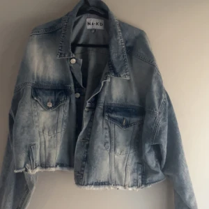 Blå croppad jeansjacka från NA-KD - Säljer en snygg blå croppad jeansjacka från NA-KD. Jackan har en cool, sliten look med råa kanter och knappar framtill. Perfekt för en avslappnad stil.