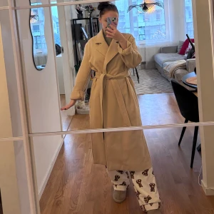 Beige kappa från H&M - Snygg beige kappa från H&M med knytskärp i midjan. Perfekt för höst och vår, med en klassisk design och långa ärmar. Kappan har en stilren look som passar till många olika outfits.