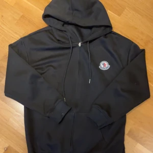 Svart zip up hoodie  - Säljer en stilren svart hoodie med dragkedja och huva. Tröjan har ett broderat märke på bröstet och långa ärmar. Perfekt för en avslappnad look. Är m men sitter som S. Kan sänka vid snabb affär. TAR EMOT BYTEN