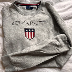 Grå sweatshirt från GANT - Säljer en stilren grå sweatshirt från GANT med broderad logotyp och emblem på bröstet. Tröjan har långa ärmar och rund halsringning. Perfekt för en avslappnad stil.
