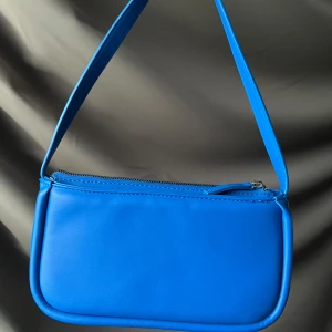 Blå axelväska i syntet - Snygg blå axelväska i syntetmaterial med en enkel och stilren design. Väskan har en praktisk dragkedja upptill och har även ett extra avtagbart långt band för bekväm passform. Perfekt för att bära dina nödvändigheter med stil.