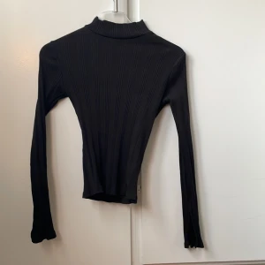 Svart ribbad polotröja - Snygg svart ribbad polotröja med långa ärmar. Perfekt för en stilren look och passar bra till både jeans och kjol. Tröjan har en tight passform som framhäver figuren.