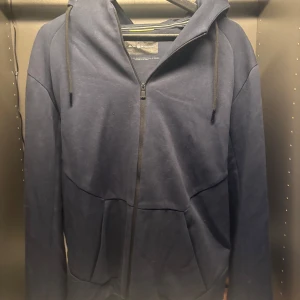 Mörkblå huvtröja från Sail Racing - Säljer en mörkblå huvtröja från Sail Racing i storlek L. Tröjan har en dragkedja framtill och en justerbar huva med dragsko. Den har ett stilrent märke på ärmen och är perfekt för en avslappnad stil.