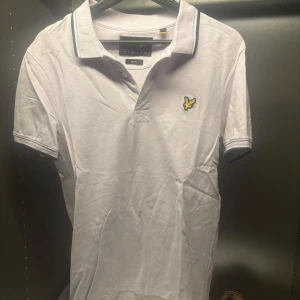 Vit pikétröja från Lyle & Scott - Säljer en stilren vit pikétröja från Lyle & Scott med klassisk krage och knappslå. Tröjan har en broderad logotyp på bröstet och kontrasterande ränder på kragen och ärmsluten. Perfekt för en avslappnad men ändå snygg look.