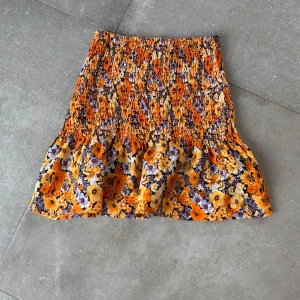 Blommig kortkjol i orange och lila - Säljer en färgglad kortkjol med blommigt mönster i orange, lila och gult. Kjolen har en volangdetalj längst ner som ger en lekfull touch. Perfekt för sommarens varma dagar!