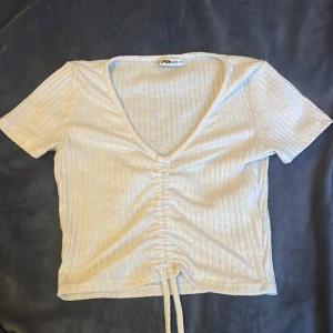 Vit ribbad topp från new yorker - Säljer en vit ribbad topp från New Yorker med korta ärmar och v-ringning. Toppen har en snygg knytdetalj framtill som ger en fin passform. Perfekt för en stilren look.