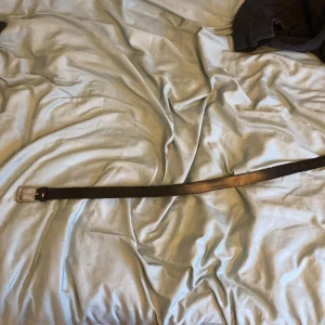 Brunt skärp med silverfärgat spänne - Säljer ett stilrent brunt skärp från Tommy Hilfiger med ett silverfärgat spänne. Skärpet har en klassisk design med flera hål för justering och en liten logotypdetalj nära spännet. Perfekt för att ge en elegant touch till din outfit.