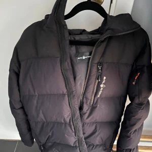 Svart dunjacka från SailRacing - Säljer en svart dunjacka från SailRacing med dragkedja och en liten logga på ärmen. Jackan har en quiltad design och en huva för extra värme. Perfekt för kyliga dagar!