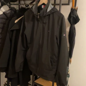 Svart vindjacka från Tommy Hilfiger - Säljer en stilren svart vindjacka från Tommy Hilfiger. Jackan har en praktisk huva och dragkedja framtill. Den är långärmad och har en liten logotyp på ärmen. Perfekt för blåsiga dagar!