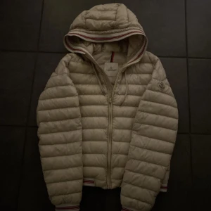 Beige dunjacka från Moncler - Säljer en snygg beige dunjacka från Moncler med huva och dragkedja. Jackan har ett klassiskt quiltat mönster och Moncler-logga på ärmen. Perfekt för kyliga dagar med stil. Tillkommer defekter som visas på bild 4,5! OBS början på dragkedjan är lite söndrig men funkar ändå, syns inte när du har på den!!!Pris ej hugget i sten🪨Skriv vid minsta lilla intresse!! Mvh John💛💛🐝🐝