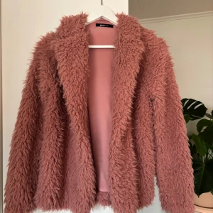 Rosa teddyjacka från Gina Tricot - Mysig rosa teddyjacka från Gina Tricot med långärmad design. Perfekt för att hålla sig varm och stilren under kyliga dagar. Jackan har en fluffig textur som ger en trendig look.