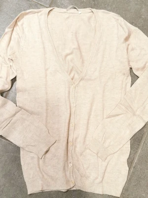 Snygg beige tunn kofta med knappar - Ljusbeige tunnstickad kofta med djup V-ringning och knäppning framtill. Långa ärmar och ribbade muddar vid ärmslut och nederkant. Perfekt att slänga över en topp för en enkel och clean look.