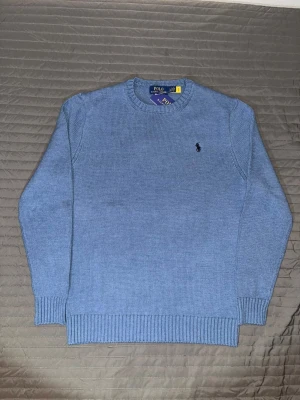 Blå stickad tröja från Polo Ralph Lauren - Snygg ljusblå stickad tröja från Polo Ralph Lauren med rund halsringning och diskret broderad logga på bröstet. Tröjan har ribbade muddar vid ärmslut och nederkant. Perfekt för dig som gillar klassisk och stilren design.