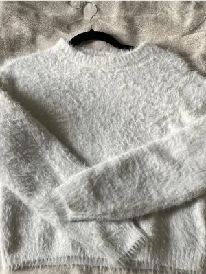 Vit fluffig stickad tröja - Mysig vit stickad tröja med fluffig yta. Tröjan har långa ärmar och ribbade muddar vid ärmslut och nederkant. Perfekt för kalla dagar när du vill vara både varm och stilren. Lite sliten efter tvätt men fortfarande skön. Ej använt på väldigt länge. Ej bra pris,, skriv till mig