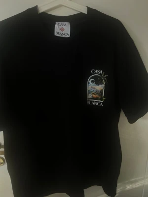 Svart Casa Blanca t-shirt med tryck - Säljer en svart t-shirt från Casa Blanca med färgglatt tryck av stadsmotiv och palm både fram och bak. T-shirten har rund hals och korta ärmar. Perfekt för dig som gillar streetwear och vill sticka ut med en unik design.