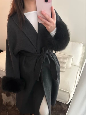Mörkgrå kappa med fuskpälsdetaljer - Stilren mörkgrå kappa med stora slag och knytskärp i midjan. Ärmsluten har lyxiga detaljer i svart fuskpäls som ger en trendig touch. Perfekt för dig som vill ha en elegant och mysig look under kallare dagar.