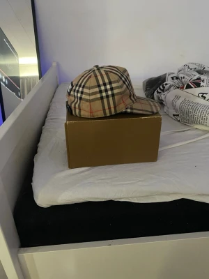 Burberry beige rutig keps - Säljer en klassisk beige keps från Burberry med det ikoniska rutmönstret i svart, vitt och rött. Kepsen har justerbar rem bak och Burberry-logga på baksidan. Tillverkad i bomull och levereras med originalkartong.