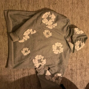 Grå denim tears  - Säljer en grå hoodie med huva och stora vita blommönster över hela tröjan. Hoodien har en klassisk känguruficka framtill och ribbade muddar vid ärmslut och nederkant. Perfekt för dig som gillar en avslappnad och trendig stil.Tröjan är i helt nytt skick och aldrig använd 