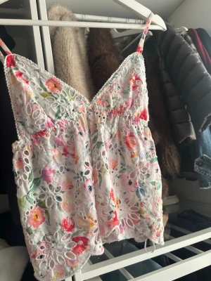 Blommig broderad linnetopp - Söt linnetopp med tunna axelband och v-ringning. Toppen är vit med färgglatt blommigt mönster och har broderade detaljer samt hålmönster. Perfekt för varma dagar och ger en romantisk vibe.