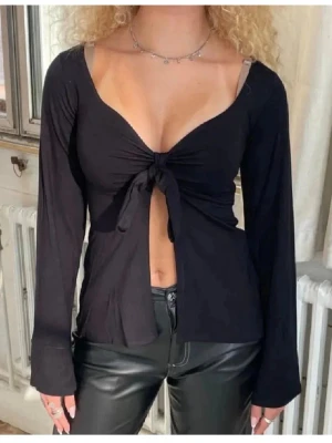 Svart långärmad topp med knyt - Trendig svart långärmad topp från Design Si med djup v-ringning och knyt framtill. Toppen har en croppad passform och är tillverkad i mjukt material som känns skönt mot huden. Perfekt för dig som vill ha en stilren men ändå edgy look.