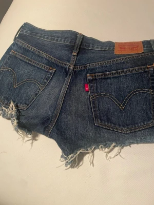Levi's jeansshorts med fransig kant - Säljer ett par klassiska blå jeansshorts från Levi's med fransig, rå kant och femficksmodell. Shortsen har normal passform och är tillverkade i slitstark bomull. Perfekta för sommaren och har den ikoniska Levi's-detaljen på bakfickan.