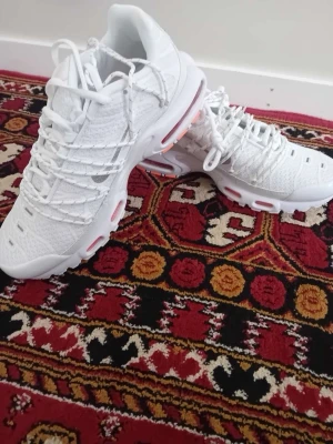 Vita Nike Air Max Plus sneakers - Snygga vita Nike Air Max Plus sneakers med mesh- och syntetmaterial, ikoniska vågiga detaljer och synliga Air-enheter i sulan med orange inslag. Skorna har en sportig look och är perfekta för dig som gillar streetwear och vill sticka ut.