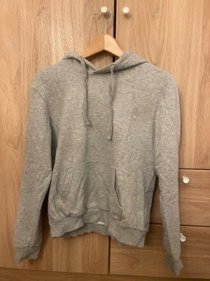 Grå basic hoodie med ficka - En enkel och stilren grå hoodie med huva och dragsnören. Hoodien har en stor magficka framtill och ribbade muddar vid ärmar och nederkant. Perfekt för chill dagar eller när du vill ha en clean look. Tillverkad i mjukt sweatshirtmaterial.