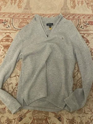 Grå hoodie från Polo Ralph Lauren - Snygg grå hoodie från Polo Ralph Lauren med liten broderad logga på bröstet. Hoodien har en avslappnad passform, V-ringad hals och långa ärmar. Tillverkad i mjukt sweatshirtmaterial som är perfekt för chill dagar.