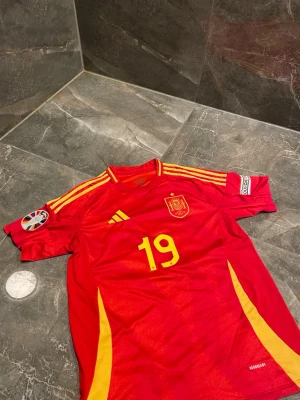 Spaniens matchtröja Adidas röd - Röd matchtröja från Adidas för Spaniens landslag med gula detaljer, nummer 19 på bröstet och officiellt landslagsemblem. Tröjan har gula ränder på axlarna, rund hals och patch på ärmen. Tillverkad i lätt och ventilerande material.