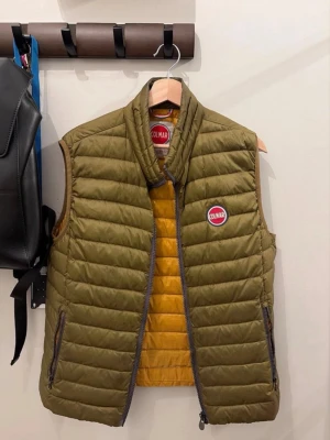 Olivgrön dunväst från Colmar - Snygg olivgrön dunväst från Colmar med quiltad design och gul insida. Västen har två dragkedjefickor, hög krage och Colmar-logga på bröstet. Perfekt för lager på lager och ger en sportig vibe till din outfit.