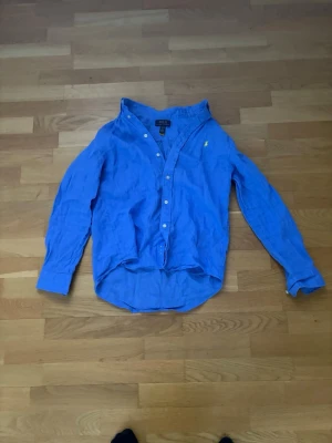 Blå skjorta från Polo Ralph Lauren - Klassisk blå skjorta från Polo Ralph Lauren med gul broderad logga på bröstet. Skjortan har lång ärm, knappar framtill och är tillverkad i bomull. Perfekt för en stilren och avslappnad look.