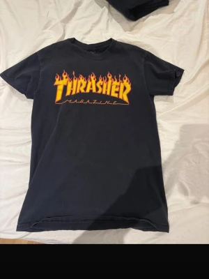 Svart Thrasher t-shirt med flames - Säljer en svart Thrasher t-shirt med den ikoniska loggan i gult och rött flames-tryck på bröstet. Klassisk passform och rund hals. Perfekt för dig som gillar streetwear och skatekultur.