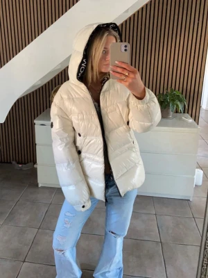 Vit dunjacka med huva från Moncler - Snygg vit dunjacka från Moncler med stor huva och svarta detaljer. Jackan har dragkedja framtill, tryckknappar och en Moncler-logga på ärmen. Perfekt för kalla vinterdagar och riktigt trendig look.