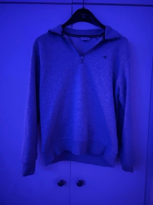 Grå halvzip hoodie från GANT - Snygg grå halvzip hoodie från GANT med broderad logga på bröstet. Tillverkad i mjukt sweatshirtmaterial och har en klassisk krage utan huva. Perfekt för en chill och stilren look.