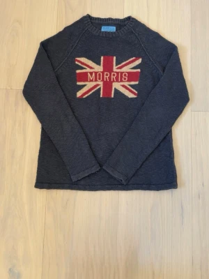 Mörkblå stickad tröja Morris - Mörkblå stickad tröja från Morris med Union Jack-flagga och MORRIS-text på bröstet. Tröjan har rund halsringning och långa ärmar. Perfekt för dig som gillar klassisk brittisk stil och vill ha något bekvämt och snyggt till vardags. Tyvärr så har den några fläckar på framsidan