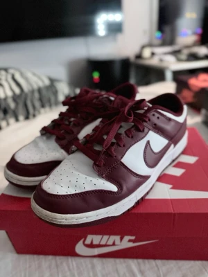 Nike Dunk Low Bordeaux sneakers - Snygga Nike Dunk Low sneakers i färgen Bordeaux och vitt. Skorna har klassisk låg siluett, vit tåbox och sidor med vinröda detaljer och swoosh. Tillverkade i läder med matchande vinröda skosnören och vit yttersula. Perfekta för dig som gillar streetwear och stilrena färgkombinationer.