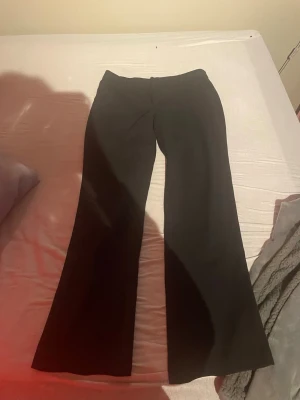 Svarta kostymbyxor Gina Tricot - Snygga svarta kostymbyxor från Gina Tricot i storlek 42. Byxorna har en klassisk rak passform med lätt utsvängda ben och hög midja. Perfekta för en stilren look. Materialet känns mjukt och följsamt, vilket gör dem bekväma att bära hela dagen.