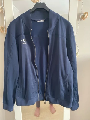 Mörkblå. Träningen jackafrån Umbro - Mörkblå  träning  jacka  från Umbro med dragkedja framtill och två sidofickor. Klassisk sportig look med Umbro-logga på bröstet. Tillverkad i mjukt material och har ribbade muddar vid ärmslut och nederkant. Perfekt för en avslappnad och sportig stil.