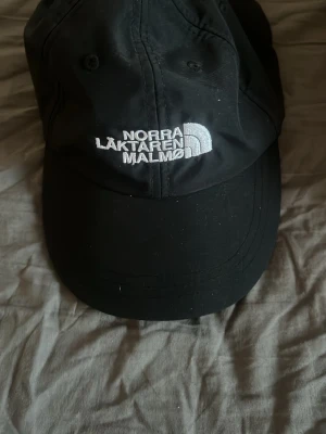 Svart keps från The North Face - Snygg svart keps med böjd skärm från The North Face. Framtill finns vit brodyr med texten 'Norra Läktaren Malmö' och den klassiska loggan. Kepsen har ventilerande hål och justerbar passform bak. Perfekt för dig som vill ha en stilren och sportig look.