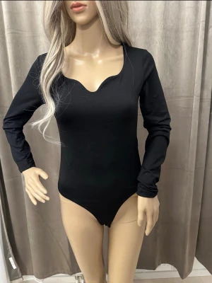 Svart långärmad body med hjärtformad urringning - Snygg svart body med långa ärmar och unik hjärtformad urringning. Tillverkad i mjukt stretchigt material som sitter skönt mot kroppen. Perfekt att styla till både jeans och kjol för en trendig look.