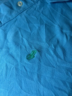 Blå pikétröja från Polo Ralph Lauren - Säljer en klassisk blå pikétröja från Polo Ralph Lauren med krage och knappar framtill. Tröjan har korta ärmar och är tillverkad i bomull, perfekt för en avslappnad och stilren look. Skickar alltid inom 24 timmar!