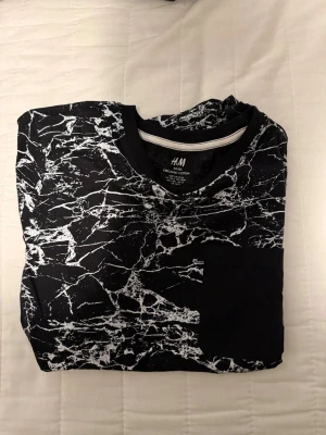 Svartvit mönstrad t-shirt från H&M - Säljer en svart t-shirt från H&M med vitt marmorinspirerat mönster. T-shirten har rund hals, korta ärmar och en svart bröstficka. Tillverkad i mjuk ekologisk bomull som känns skön mot huden. Perfekt för dig som gillar en unik och modern stil.