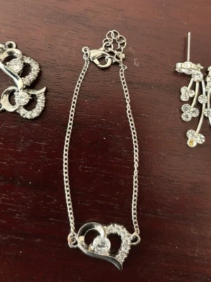 Silverfärgat armband med hjärthänge - Snyggt armband i silverfärg med ett hjärthänge dekorerat med gnistrande stenar. Kedjan är tunn och smidig. Ett par matchande örhängen till. Sen ett annat par örhängen. Perfekt för dig som gillar stilrena smycken med lite extra bling.