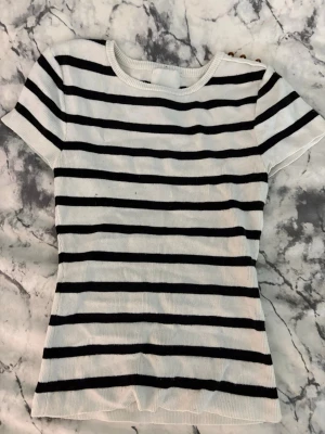 Randig ribbad topp i bomull - Snygg vit ribbad topp med svarta horisontella ränder. Kortärmad och figurnära passform, perfekt till jeans eller kjol. Tillverkad i mjuk bomull som känns skön mot huden. Enkel och stilren design som funkar till många olika looks.