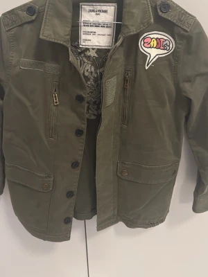 Grön overshirt från Zadig & Voltaire - Cool grön jacka från Zadig & Voltaire med broderade detaljer och patch framtill. Jackan har knappar, dragkedja och flera fickor. På ryggen finns ett stort broderat motiv med text och blommor. Perfekt för dig som vill sticka ut. Avser barn ålder 10 år