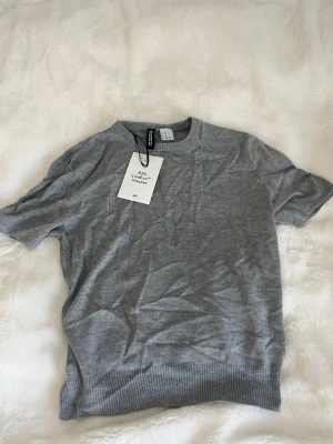 Grå kortärmad topp från H&M - En stilren grå topp från H&M i viskosblandning. Modellen har korta ärmar, rund halsringning och ribbade muddar vid ärmslut och nederkant. Perfekt att matcha med jeans eller kjol för en clean look. Liten o modellen därför säljs den. 